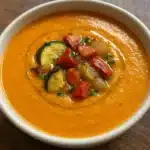 Roasted Vegetable Soup 5 A bowl of creamy orange roasted vegetable soup garnished with colorful roasted vegetables, fresh thyme, and grated Parmesan cheese, photographed from above on a rustic wooden table.